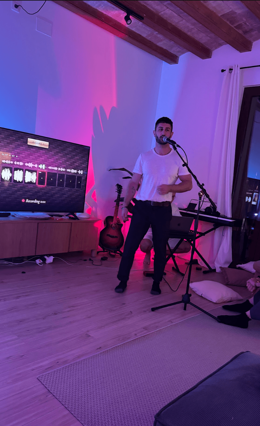 A friend at the microphone in front of the TV, waveforms on the screen, another at the keyboard in pink and blue light